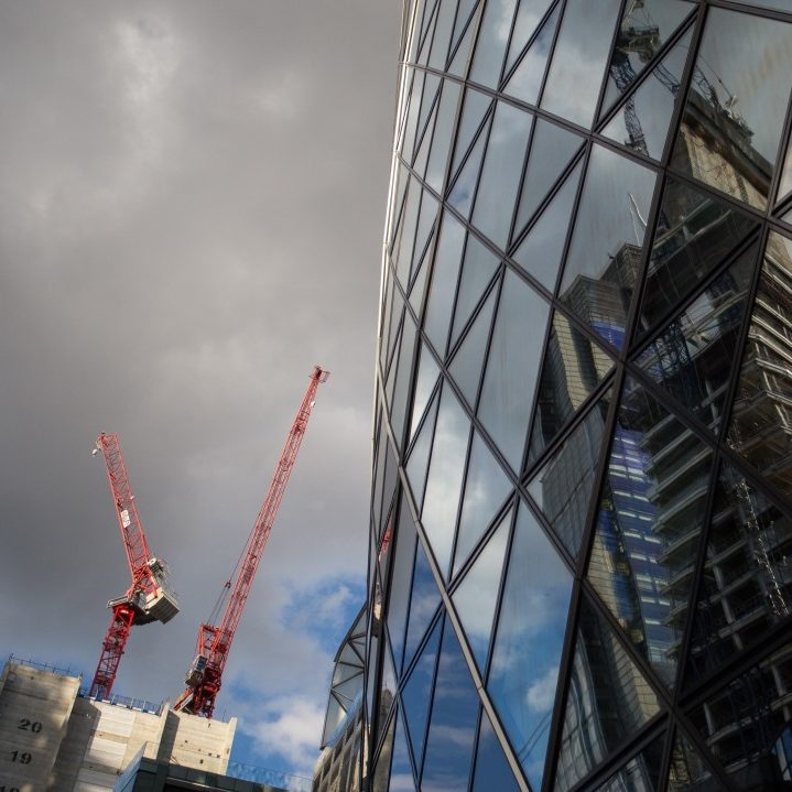Photo of London gherkin building on the right and two construction cranes on the left. 